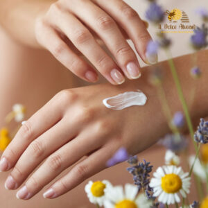 Inquadratura macro su una mano femminile che applica delicatamente un unguento cremoso, creando una sensazione di morbidezza. Sullo sfondo sfocato, piante e fiori come la camomilla o la lavanda richiamano l'origine naturale del prodotto.